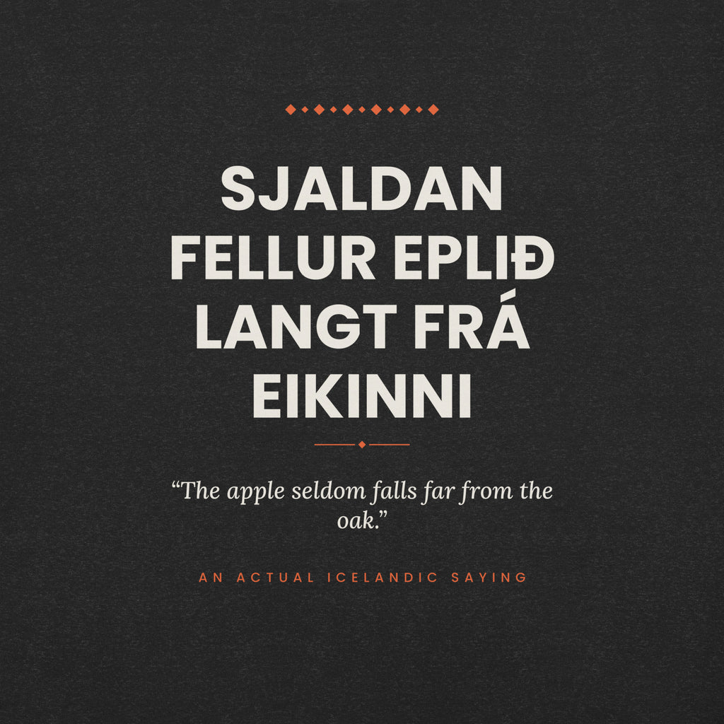 Close-up of the Sjaldan fellur eplið langt frá eikinni front print, showing cream lettering, English translation and coral accents on Black Heather fabric.