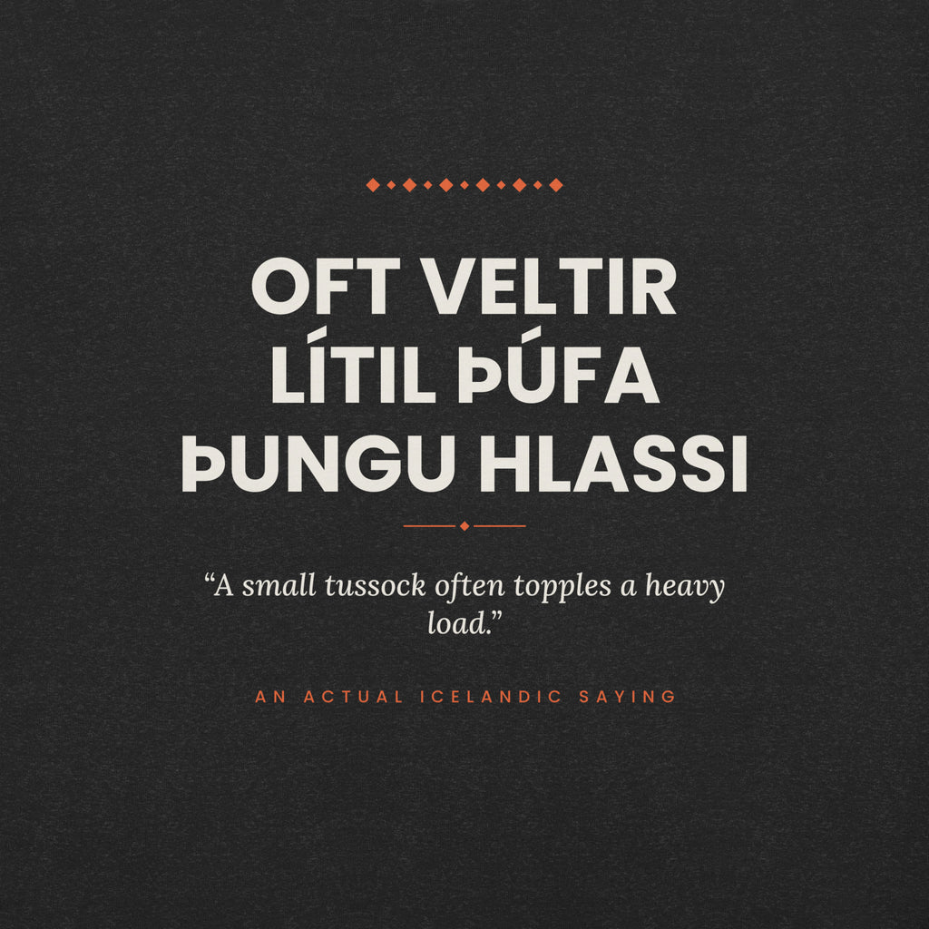 Close-up of the Oft veltir lítil þúfa þungu hlassi front print, showing cream lettering, English translation and coral accents on Black Heather fabric.