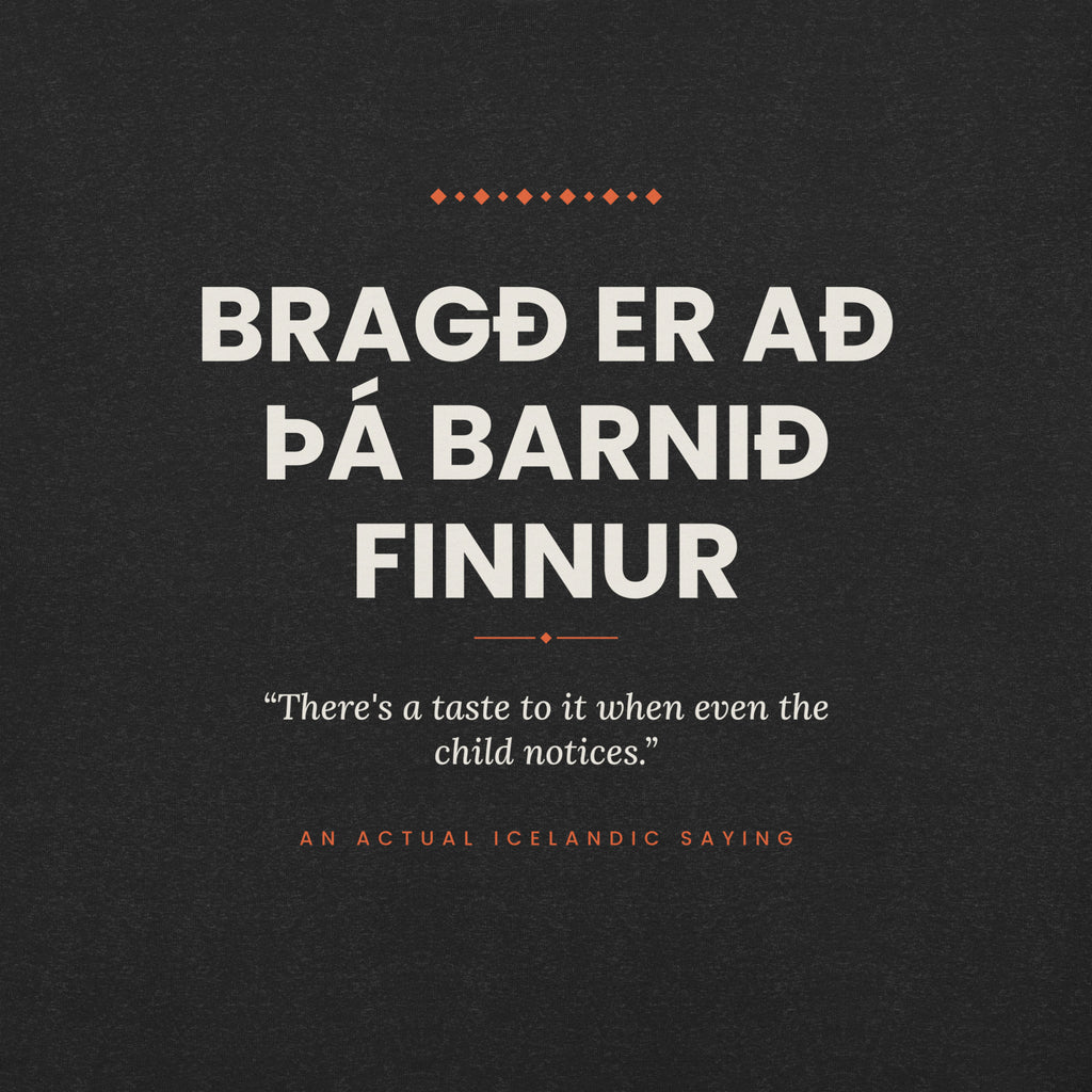Close-up of the Bragð er að þá barnið finnur front print, showing cream lettering, English translation and coral accents on Black Heather fabric.