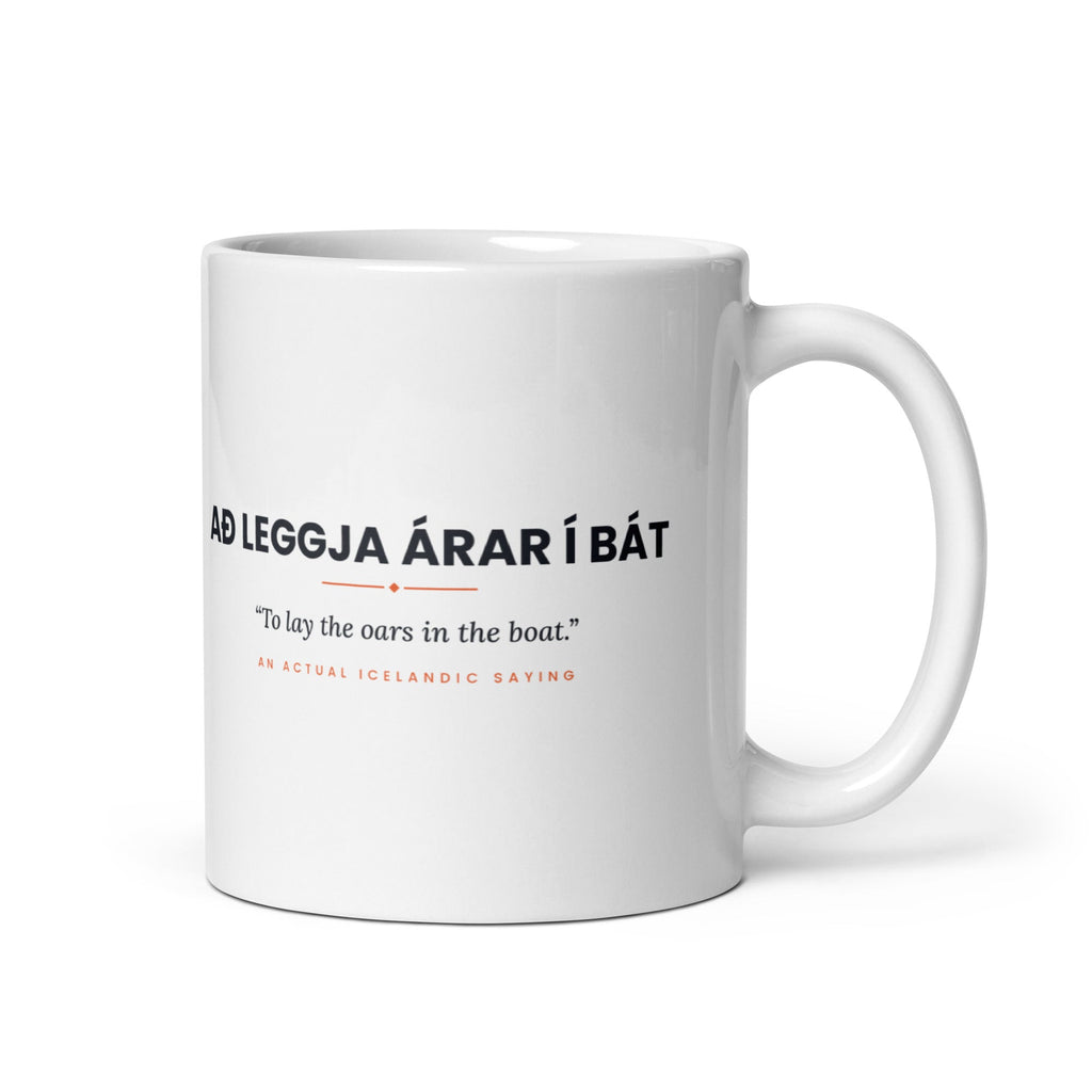 White 11 oz Að leggja árar í bát mug, handle on the right, showing black lettering, English translation and coral accents.