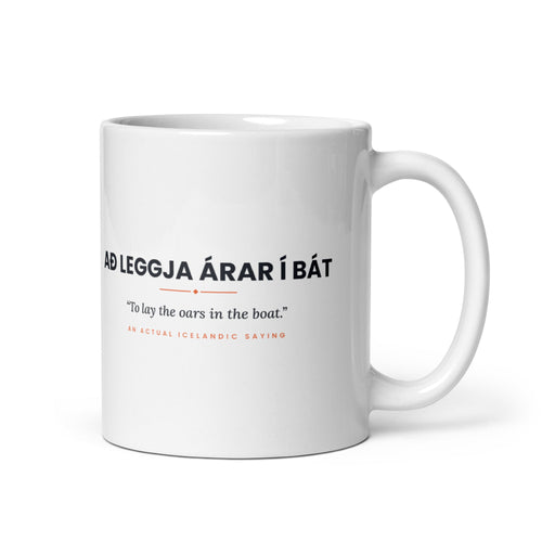 White 11 oz Að leggja árar í bát mug, handle on the right, showing black lettering, English translation and coral accents.