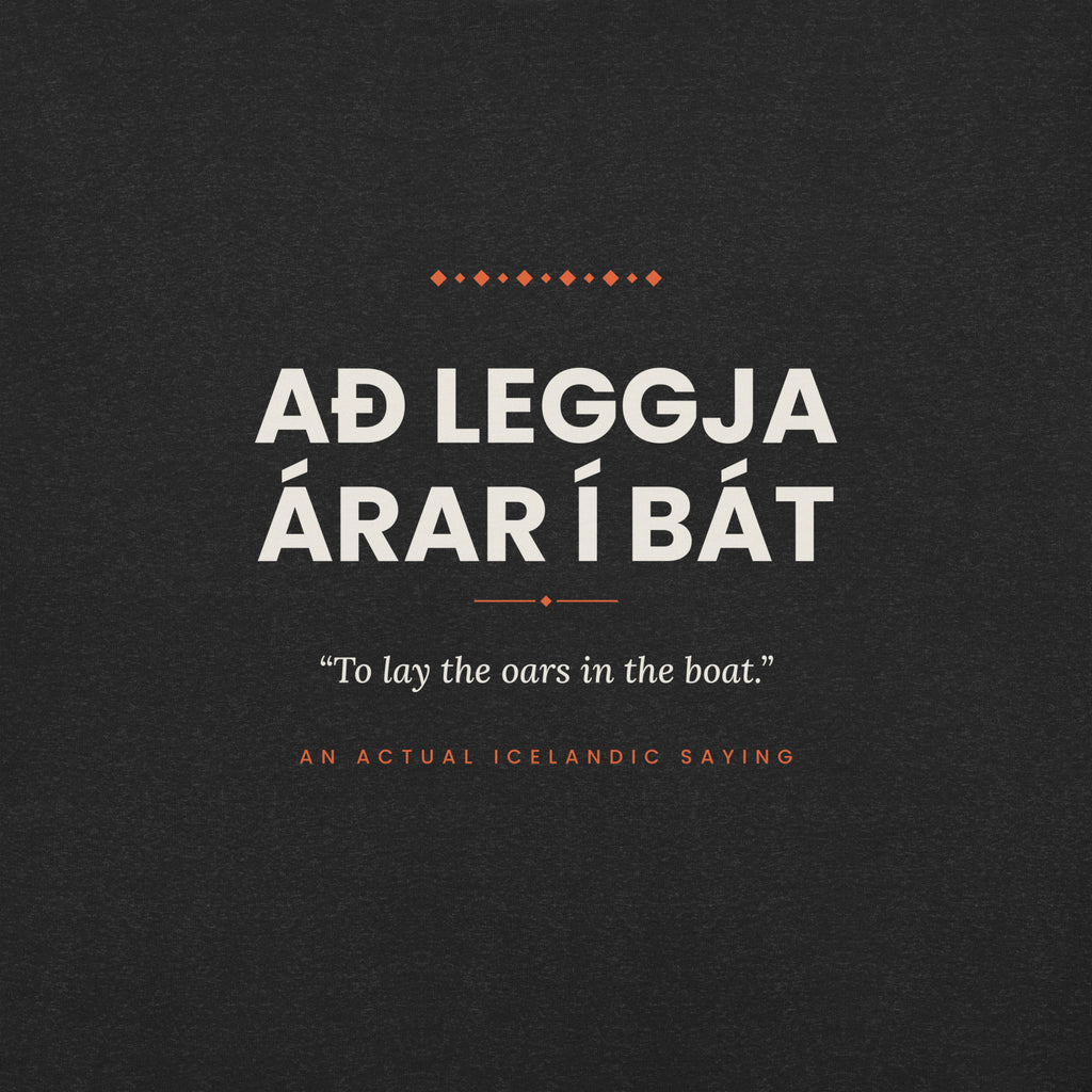 Close-up of the Að leggja árar í bát front print, showing cream lettering, English translation and coral accents on Black Heather fabric.