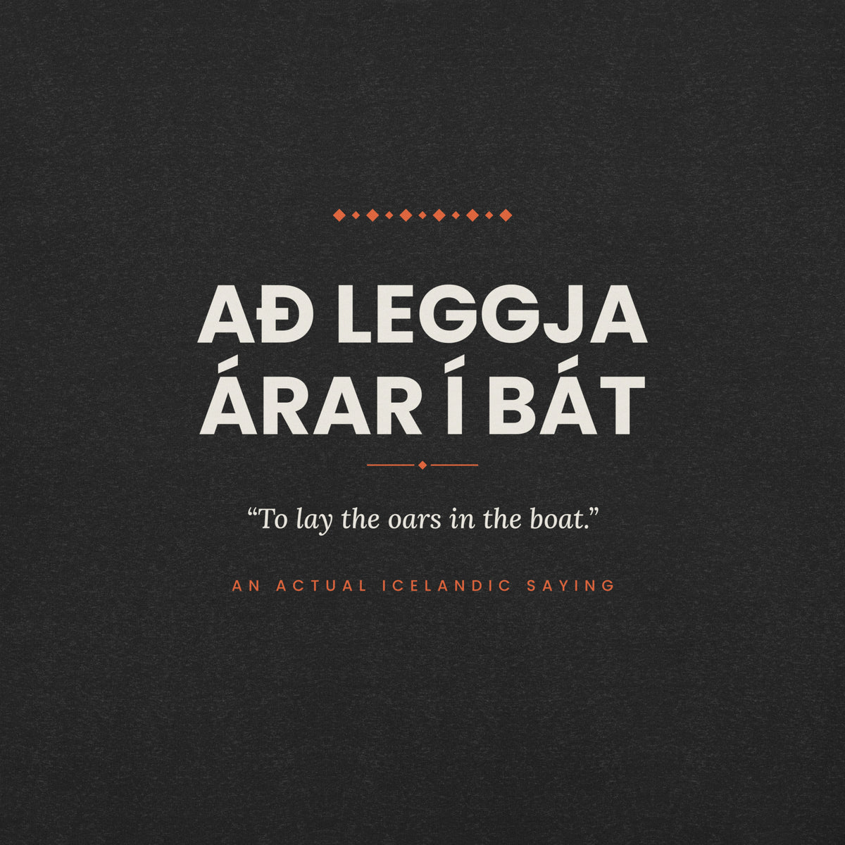Close-up of the Að leggja árar í bát front print, showing cream lettering, English translation and coral accents on Black Heather fabric.