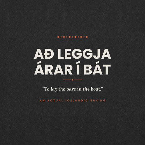 Close-up of the Að leggja árar í bát front print, showing cream lettering, English translation and coral accents on Black Heather fabric.