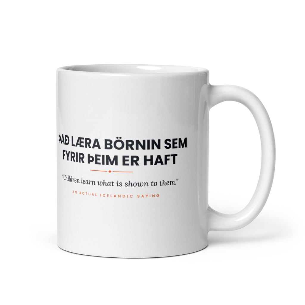 White 11 oz Það læra börnin sem fyrir þeim er haft mug, handle on the right, showing black lettering, English translation and coral accents.
