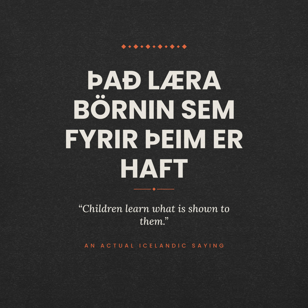 Close-up of the Það læra börnin sem fyrir þeim er haft front print, showing cream lettering, English translation and coral accents on Black Heather fabric.