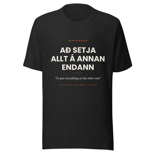 Front view of the Black Heather Að setja allt á annan endann tee, with cream lettering, English translation and coral accents.