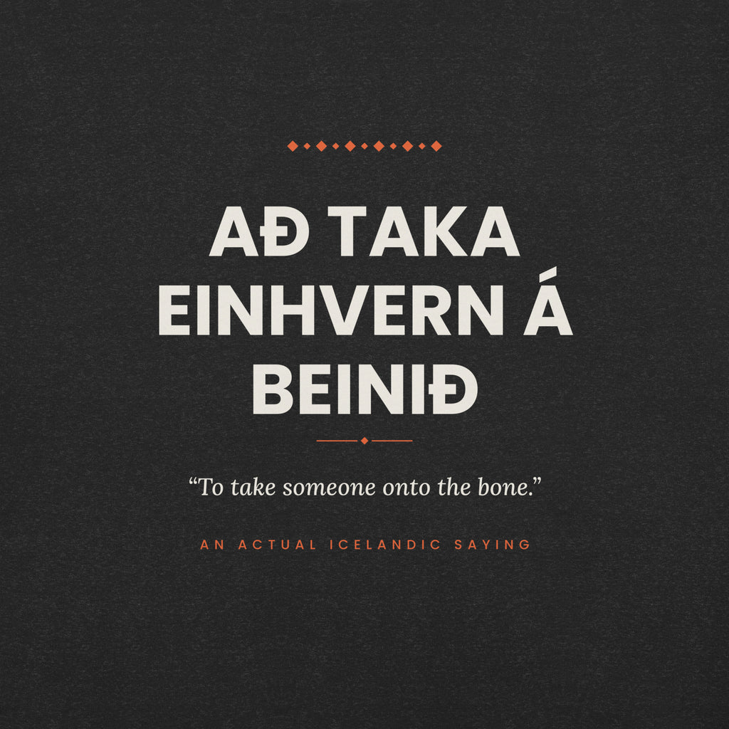 Close-up of the Að taka einhvern á beinið front print, showing cream lettering, English translation and coral accents on Black Heather fabric.