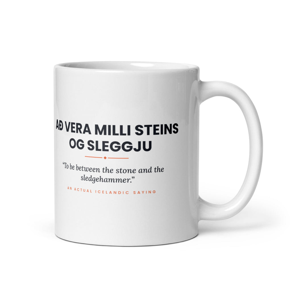 White 11 oz Að vera milli steins og sleggju mug, handle on the right, showing black lettering, English translation and coral accents.