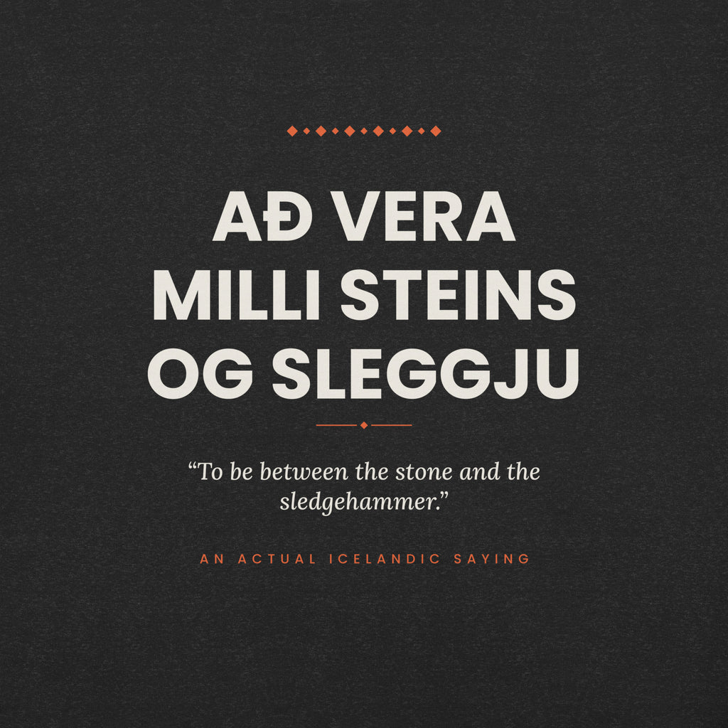 Close-up of the Að vera milli steins og sleggju front print, showing cream lettering, English translation and coral accents on Black Heather fabric.