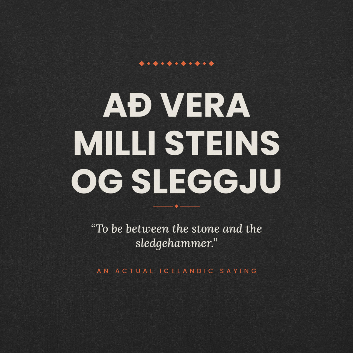Close-up of the Að vera milli steins og sleggju front print, showing cream lettering, English translation and coral accents on Black Heather fabric.
