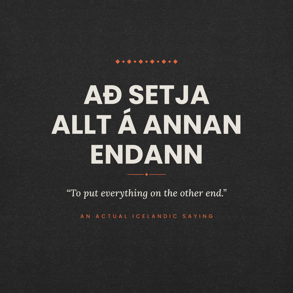 Close-up of the Að setja allt á annan endann front print, showing cream lettering, English translation and coral accents on Black Heather fabric.
