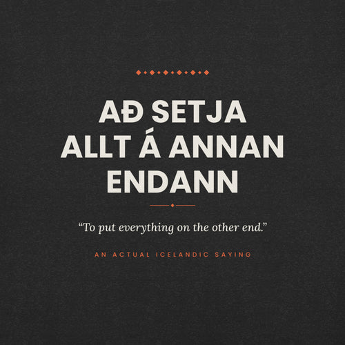 Close-up of the Að setja allt á annan endann front print, showing cream lettering, English translation and coral accents on Black Heather fabric.