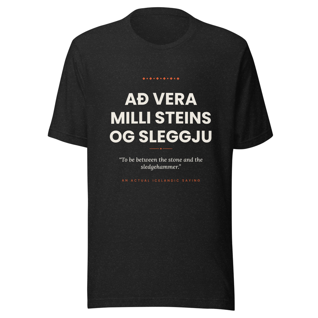 Front view of the Black Heather Að vera milli steins og sleggju tee, with cream lettering, English translation and coral accents.