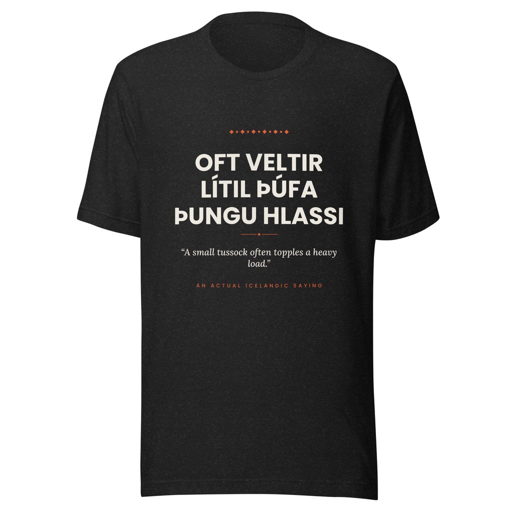 Front view of the Black Heather Oft veltir lítil þúfa þungu hlassi tee, with cream lettering, English translation and coral accents.